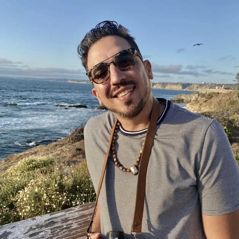 Portrait of Antonio Lopez, wearing sunglasses, a grey t-shirt, and holding a camera.