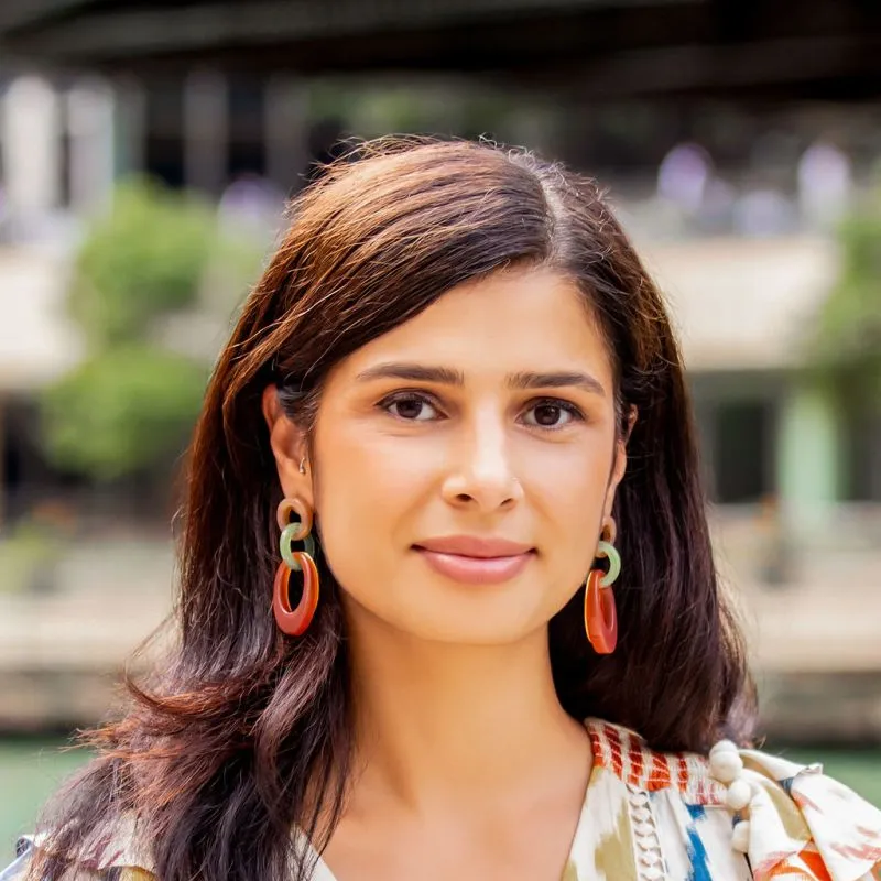 Portrait of Uzma Noormohamed with long, brown hair, colorful hoop earrings, and a white shirt with splashes of color.