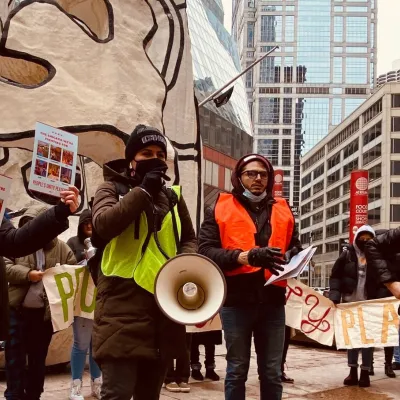 Four people at an action event stand with flyers that say "The Chicago We're Fighting For" and a megaphone. Behind them are many more people holding banners.